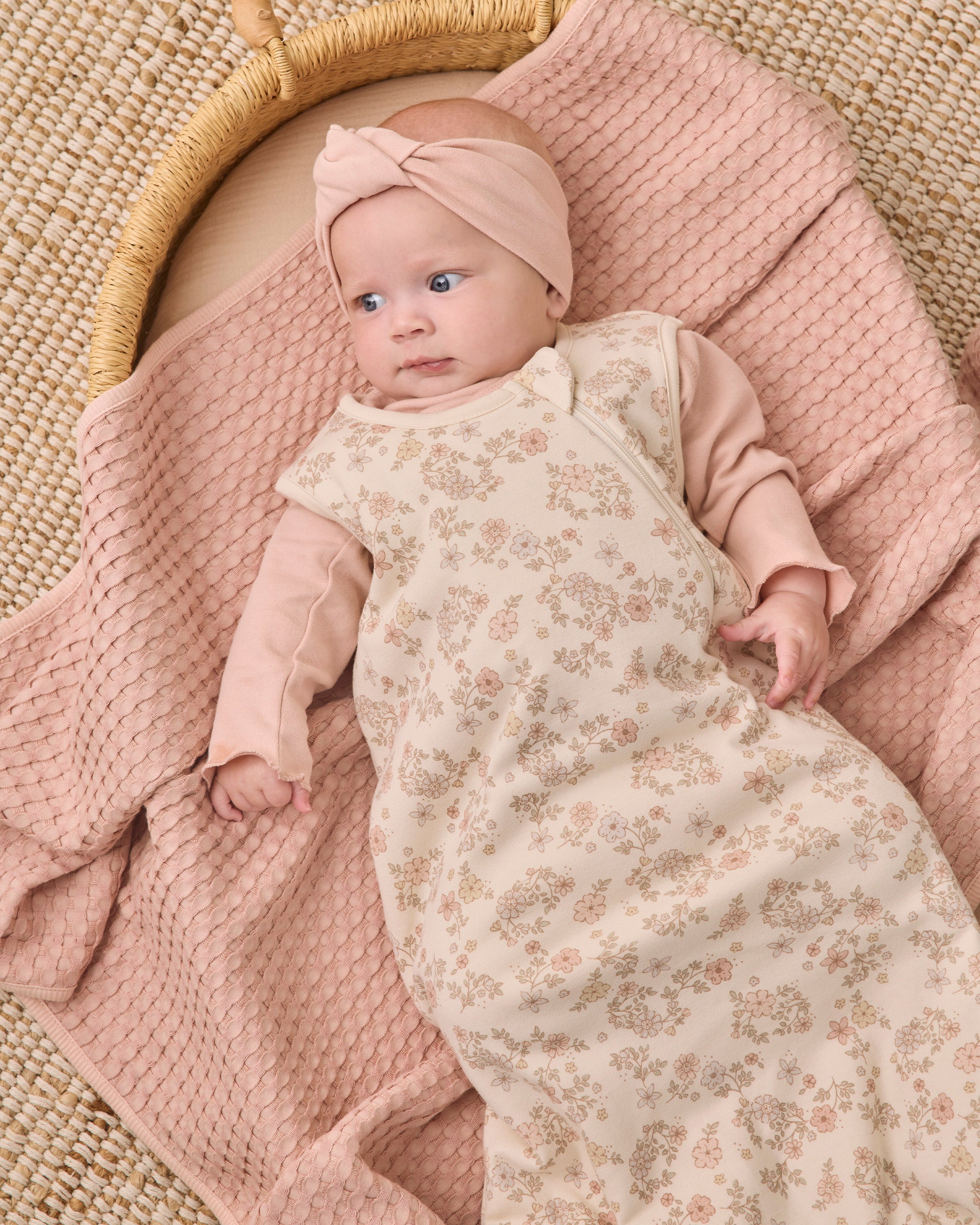 A baby lying on a woven mat under a pink textured blanket, wearing a cream floral sleep sack with long sleeves and a matching pink bow headband.