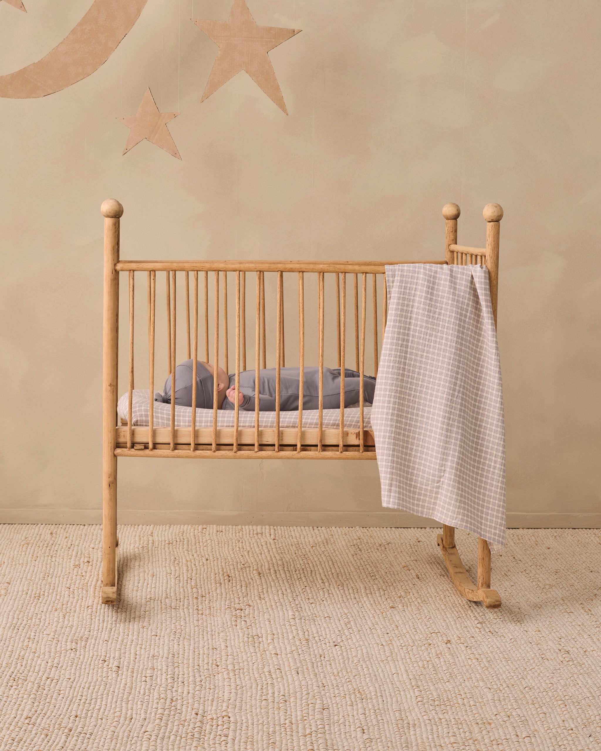A light natural wood rocking crib with vertical slats, shown in a styled nursery setting with a baby sleeping inside and a white gingham blanket draped over the side, against a beige wall with star and moon wall decals.