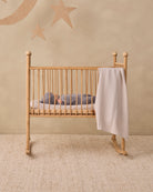A light natural wood rocking crib with vertical slats, shown in a styled nursery setting with a baby sleeping inside and a white gingham blanket draped over the side, against a beige wall with star and moon wall decals.