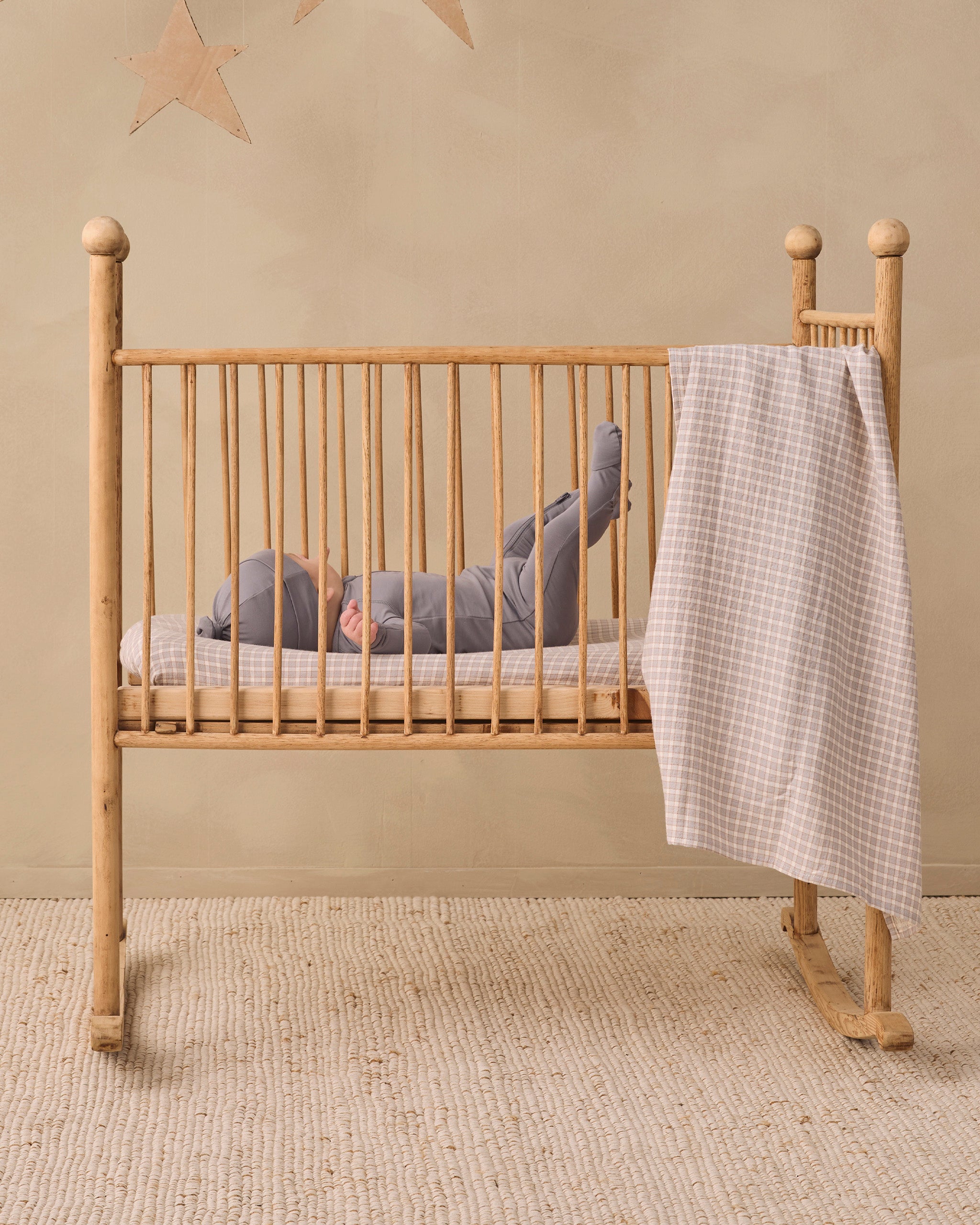 A baby in a gray outfit lies on their back inside a light natural wood rocking crib with vertical slats, with a beige and white gingham check blanket draped over the side. The crib is set on a textured beige rug against a warm beige wall with a star decoration.