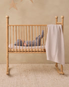 A baby in a gray outfit lies on their back inside a light natural wood rocking crib with vertical slats, with a beige and white gingham check blanket draped over the side. The crib is set on a textured beige rug against a warm beige wall with a star decoration.