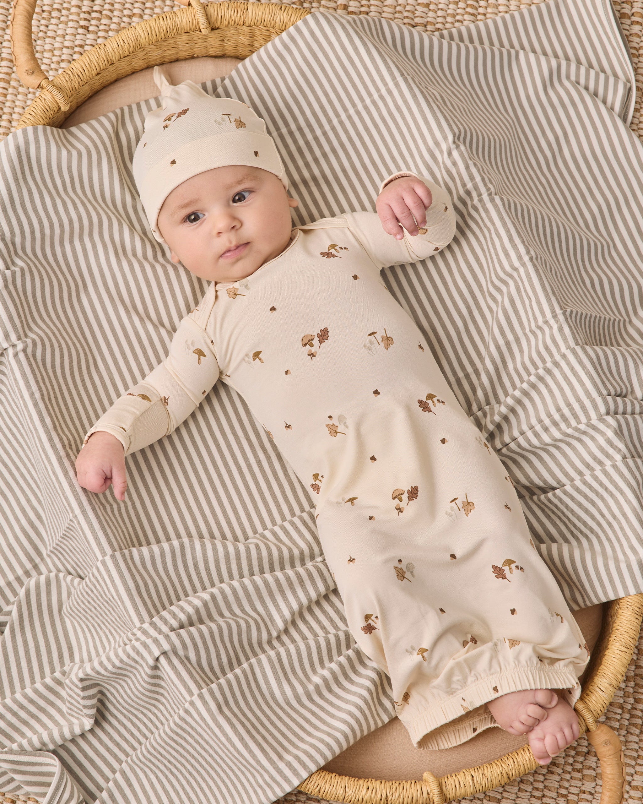 A baby lying on a striped blanket in a woven basket, wearing a cream long-sleeve romper with small brown animal prints and a matching beanie.