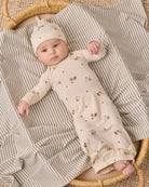 A baby lying on a striped blanket in a woven basket, wearing a cream long-sleeve romper with small brown animal prints and a matching beanie.