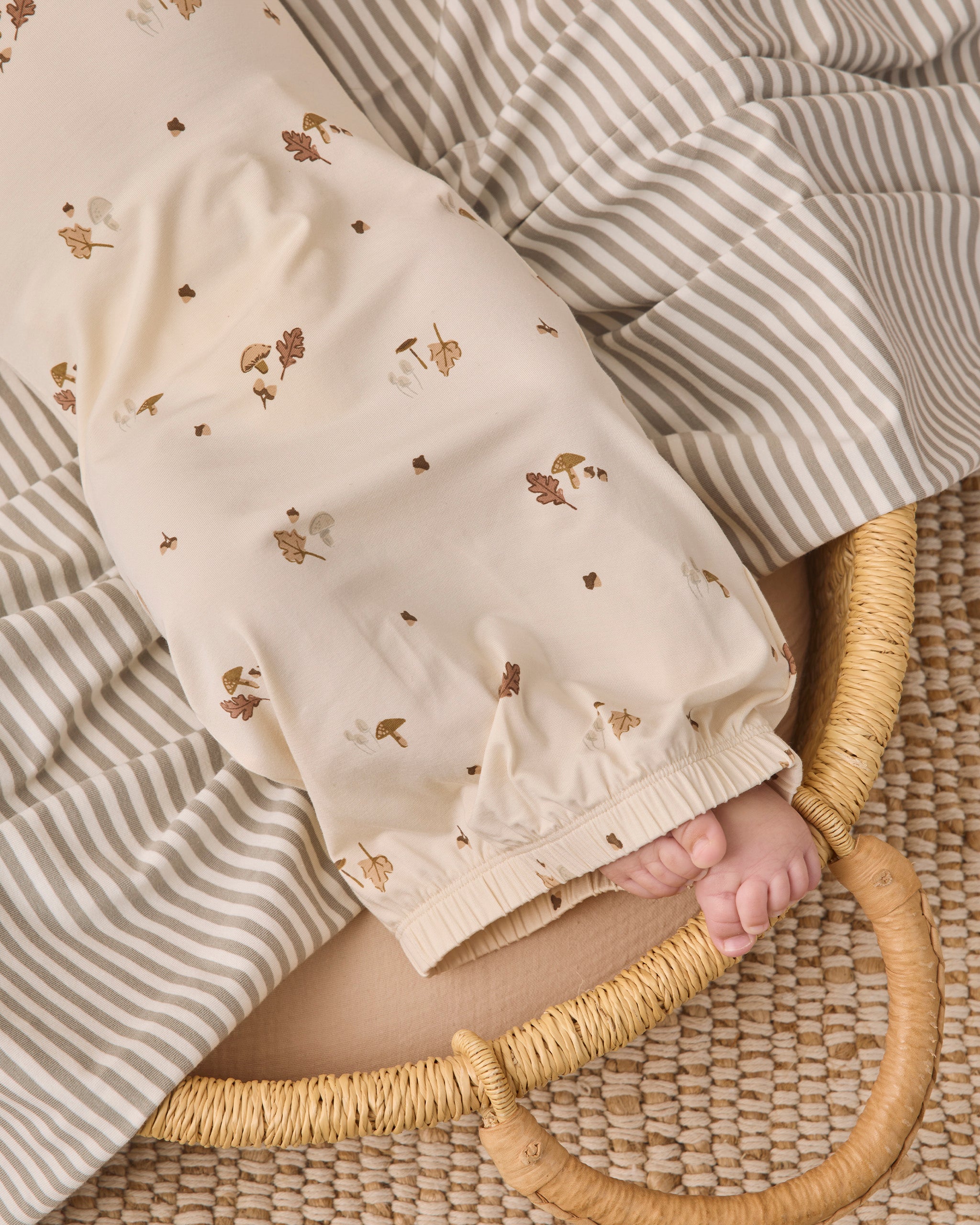 A close-up of a baby's foot and hand resting on a cream-colored blanket with a small brown and beige woodland print, tucked under a striped beige and white blanket on a woven mat.