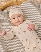 A baby lying on a striped blanket next to a woven basket, wearing a cream-colored long-sleeve bodysuit and matching beanie hat with small brown and gold woodland prints.