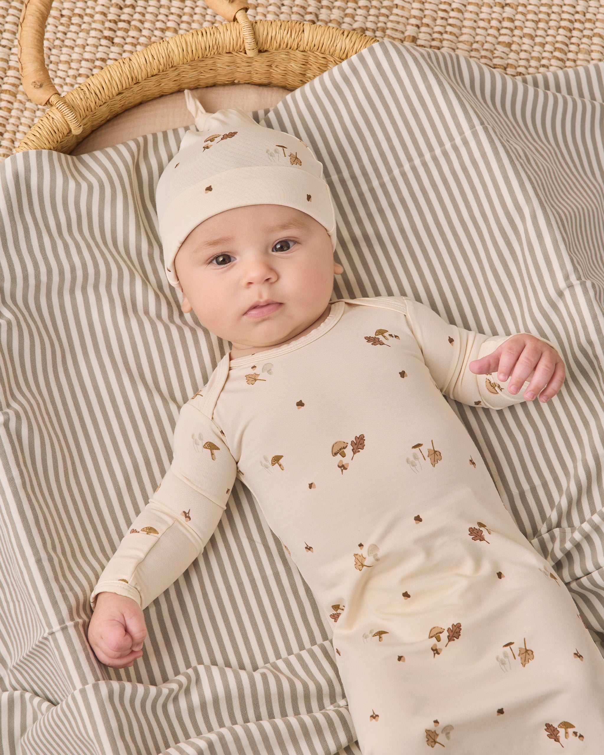 A baby lying on a striped blanket next to a woven basket, wearing a cream-colored long-sleeve bodysuit and matching hat with small brown animal prints.