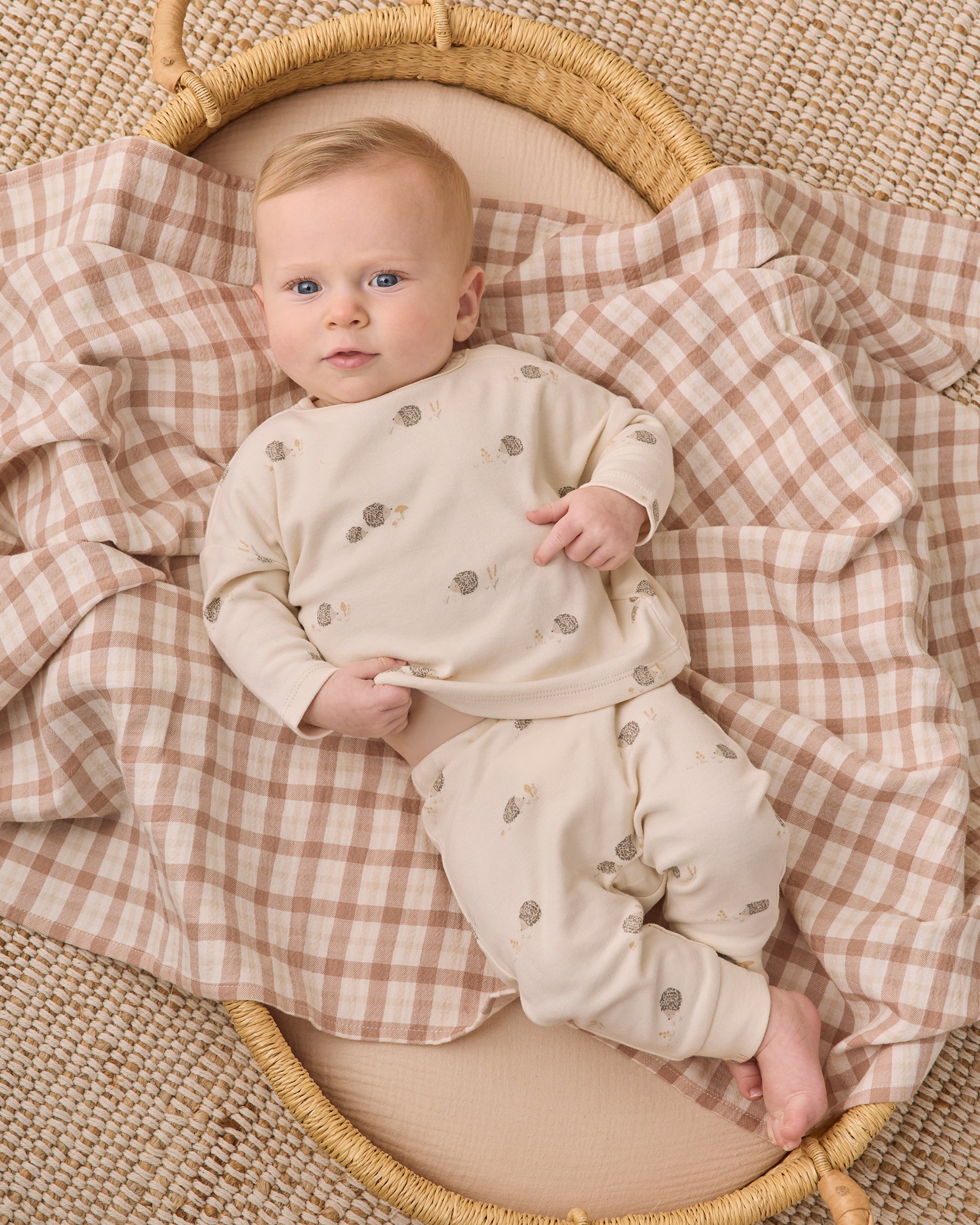 A baby lying on a woven mat, wearing a cream long-sleeve romper and matching pants with small gray tree prints, while resting on a beige and brown gingham blanket inside a wicker basket.