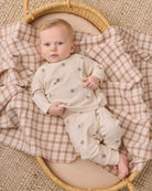 A baby lying on a woven mat, wearing a cream long-sleeve romper and matching pants with small gray tree prints, while resting on a beige and brown gingham blanket inside a wicker basket.