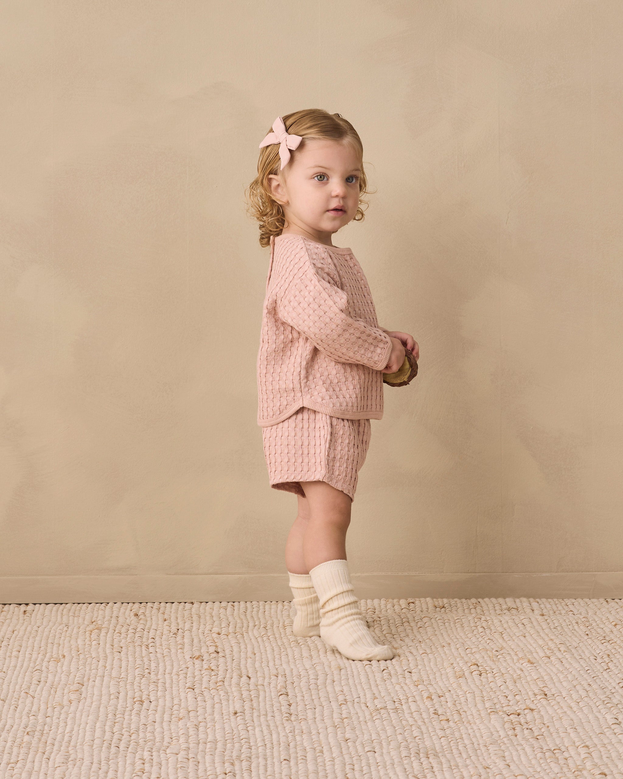 A toddler girl stands in profile wearing a matching pink textured knit outfit with a long-sleeve top and shorts, cream socks, and a pink bow in her hair, set against a warm beige textured wall and rug.