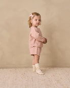 A toddler girl stands in profile wearing a matching pink textured knit outfit with a long-sleeve top and shorts, cream socks, and a pink bow in her hair, set against a warm beige textured wall and rug.