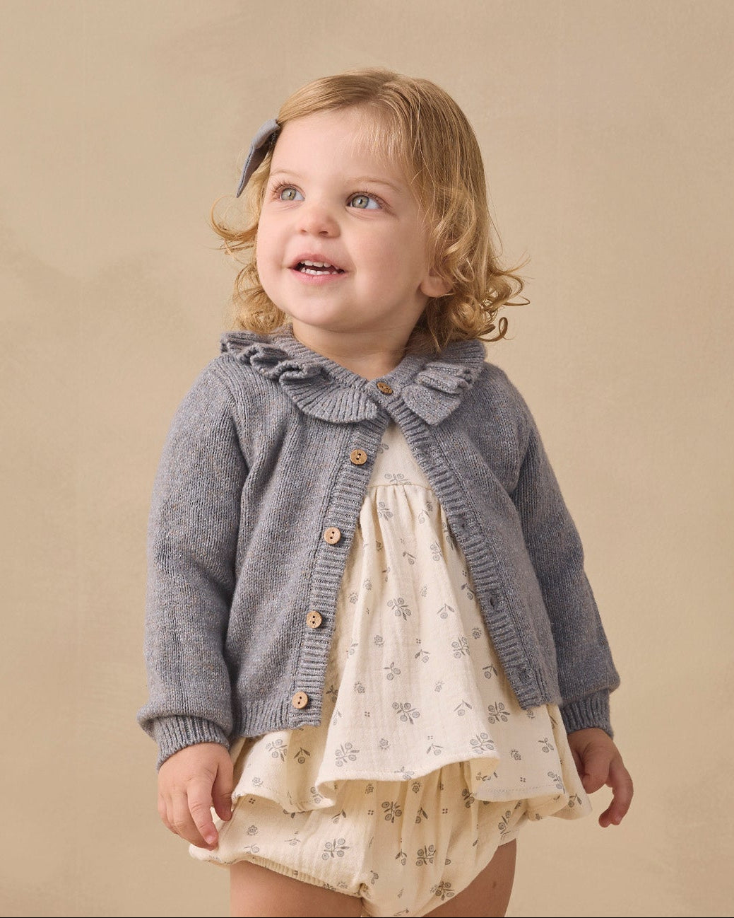 A toddler girl stands in a studio wearing a gray knit cardigan over a cream floral dress, with cream socks and tan shoes, against a warm beige backdrop.