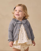 A toddler girl stands in a studio wearing a gray knit cardigan over a cream floral dress, with cream socks and tan shoes, against a warm beige backdrop.