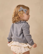 A toddler girl shown from the back wearing a gray knit cardigan, cream ruffled skirt, and a gray bow headband, set against a plain beige background.
