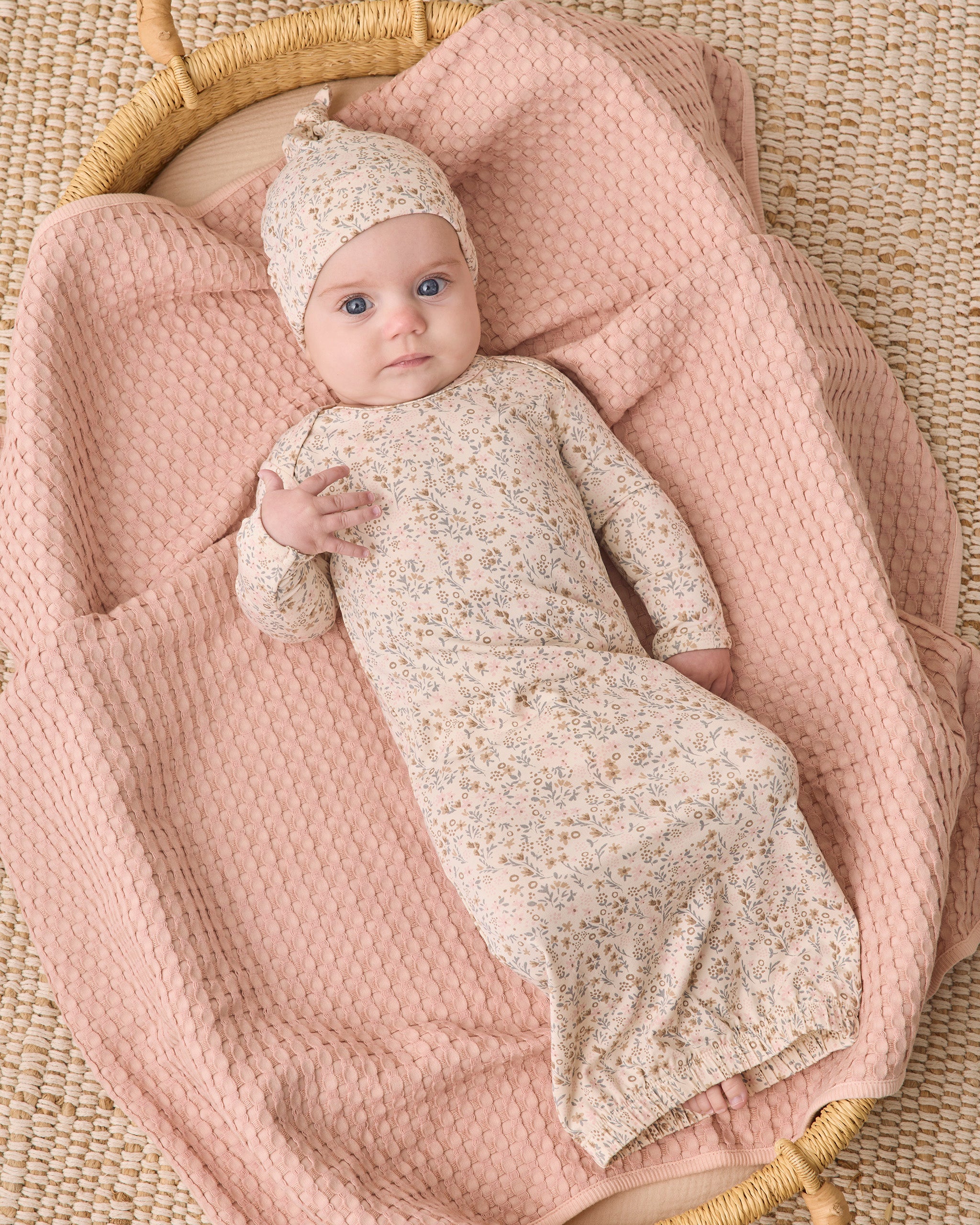 A baby wearing a cream floral long-sleeve swaddle blanket and matching knit bonnet, lying on a dusty pink textured blanket inside a woven basket.