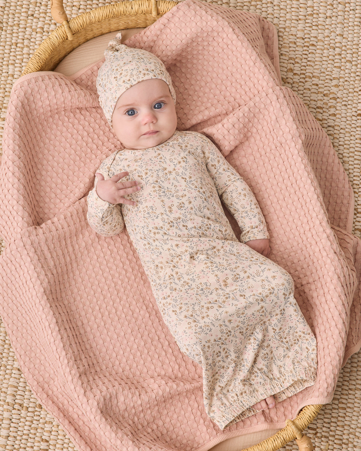 A baby wearing a cream floral long-sleeve swaddle blanket and matching knit bonnet, lying on a dusty pink textured blanket inside a woven basket.
