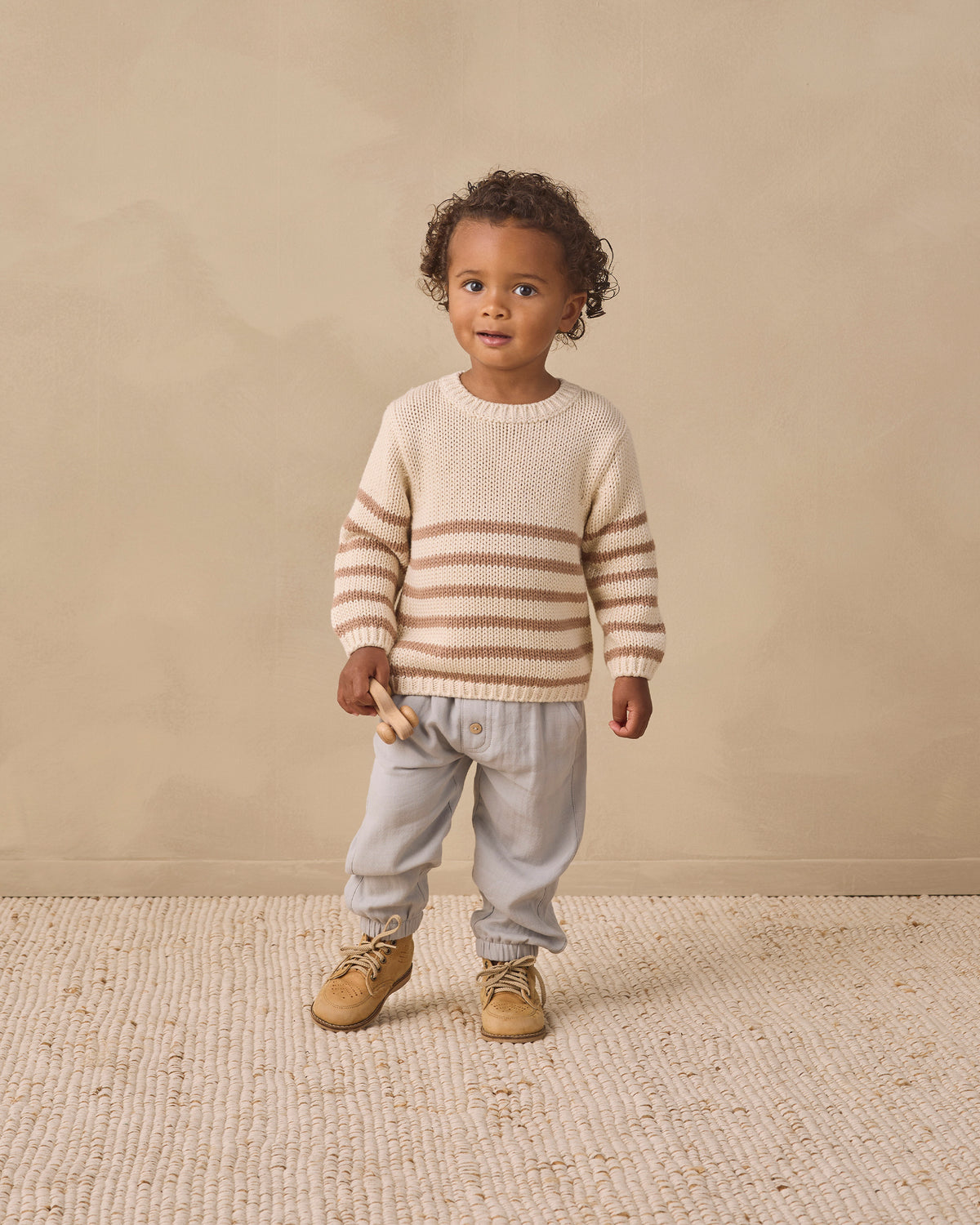 A toddler stands against a warm beige studio backdrop wearing a cream knit sweater with horizontal tan stripes, light gray pants, and tan shoes.