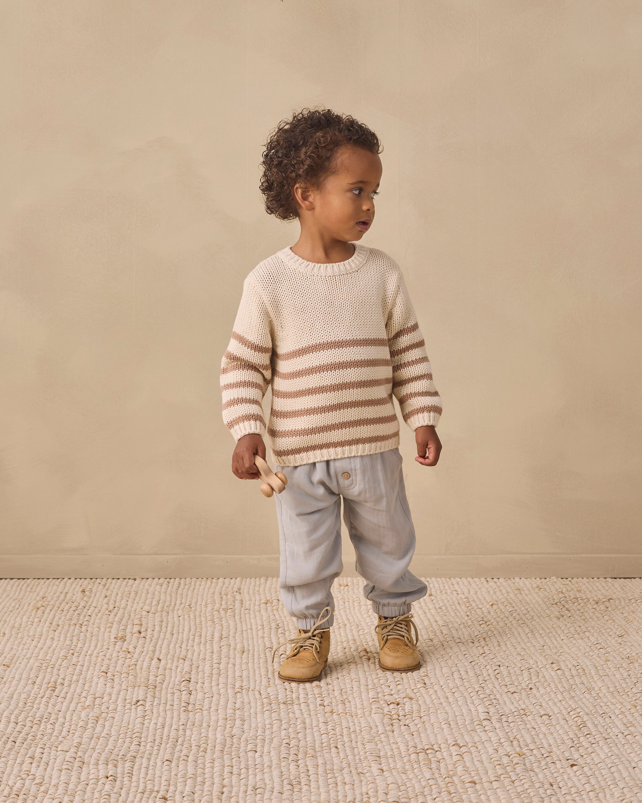 A toddler stands in profile wearing a cream knit sweater with horizontal tan stripes, light gray pants, and tan boots against a warm beige studio backdrop.