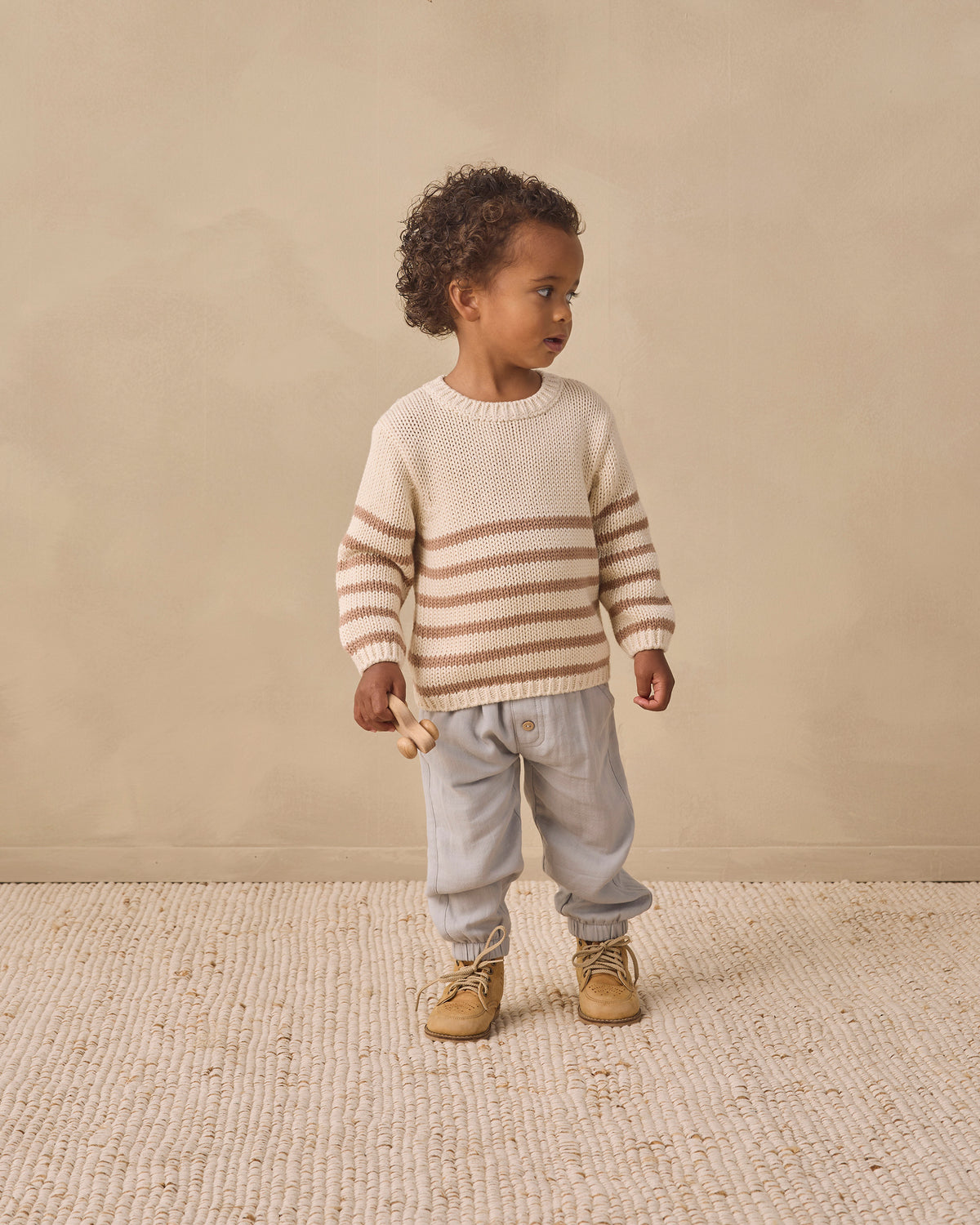 A toddler stands in profile wearing a cream knit sweater with horizontal tan stripes, light gray pants, and tan boots against a warm beige studio backdrop.