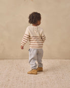 A toddler standing with their back to the camera, wearing a cream and brown striped knit sweater, light gray pants, and tan boots, set against a neutral beige textured wall and floor.