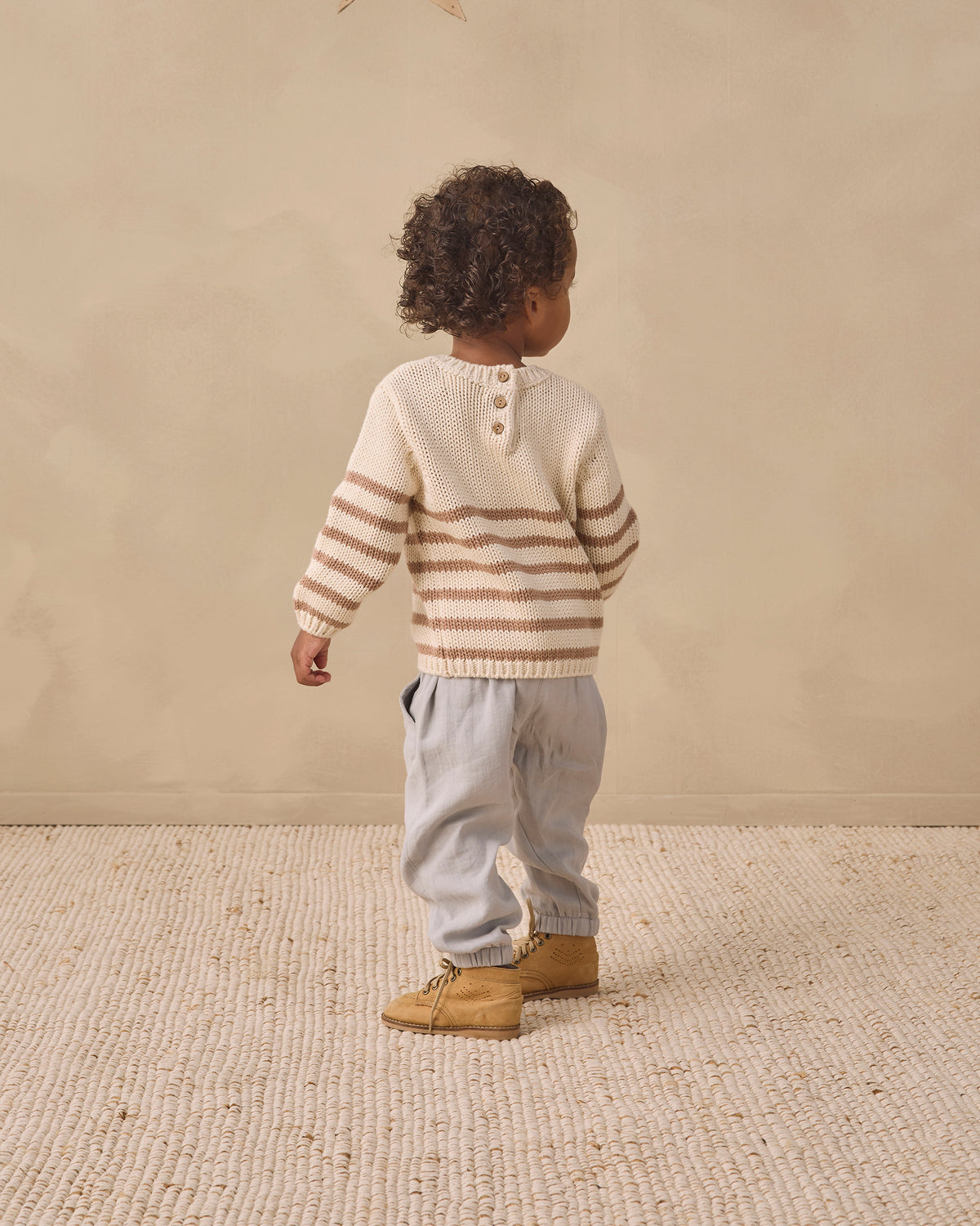 A toddler standing with their back to the camera, wearing a cream and brown striped knit sweater, light gray pants, and tan boots, set against a neutral beige textured wall and floor.