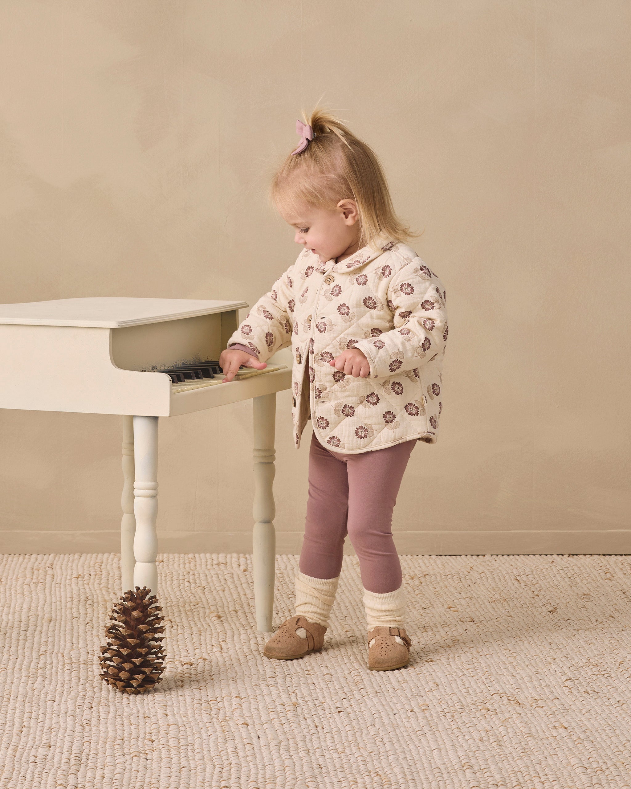 A toddler girl stands beside a small white piano in a neutral studio setting, wearing a cream quilted jacket with a brown and pink floral pattern, mauve leggings, and tan shoes.