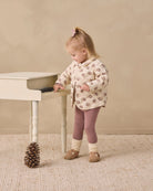 A toddler girl stands beside a small white piano in a neutral studio setting, wearing a cream quilted jacket with a brown and pink floral pattern, mauve leggings, and tan shoes.