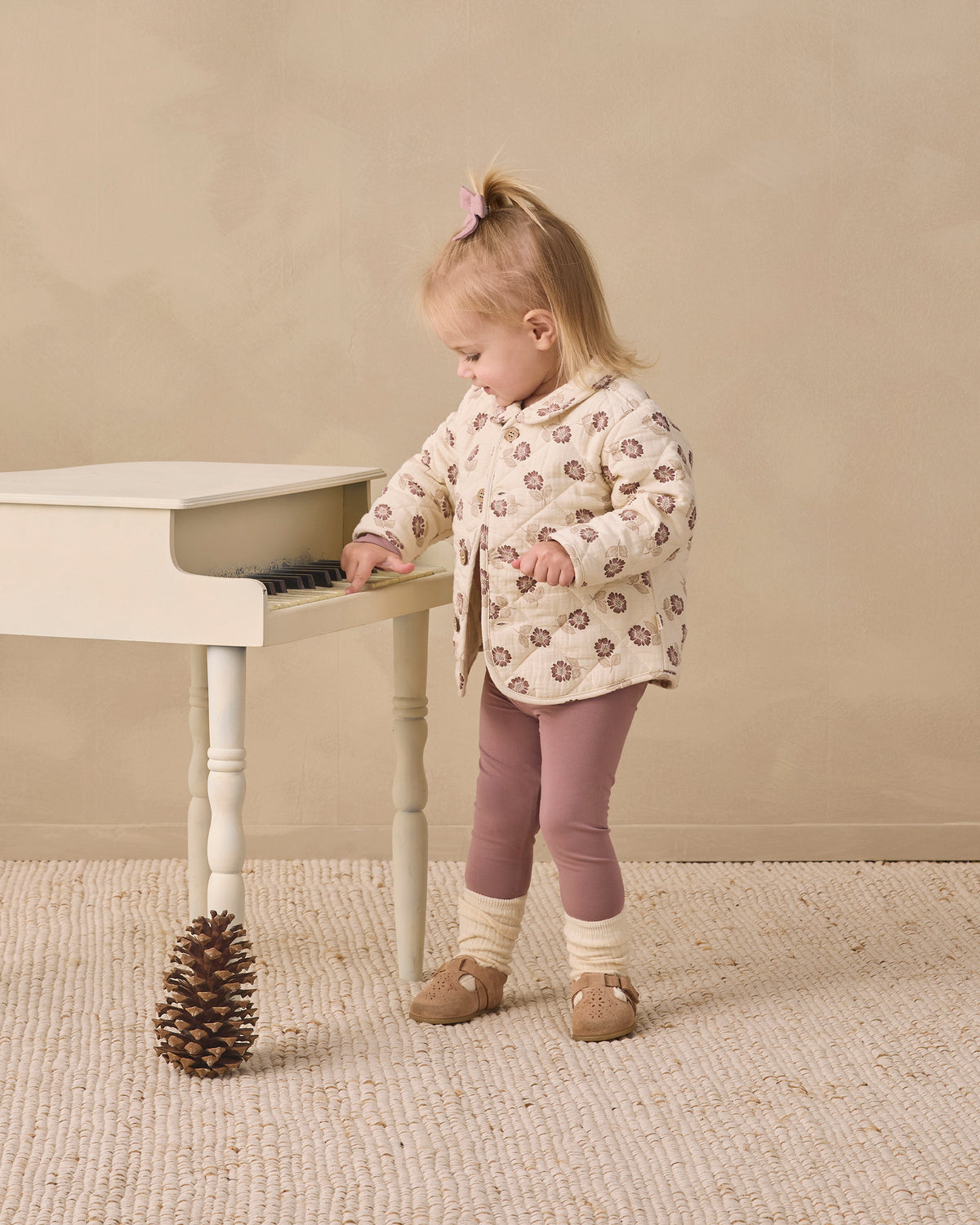 A toddler girl stands beside a small white piano in a neutral studio setting, wearing a cream quilted jacket with a brown and pink floral pattern, mauve leggings, and tan shoes.