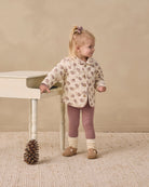 A toddler girl stands next to a small white table in a studio setting, wearing a cream-colored quilted jacket with a brown and beige pattern, mauve leggings, and tan shoes.