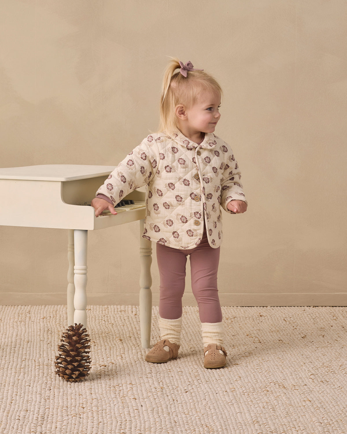 A toddler girl stands next to a small white table in a studio setting, wearing a cream-colored quilted jacket with a brown and beige pattern, mauve leggings, and tan shoes.