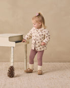 A toddler girl stands next to a small white table in a studio setting, wearing a cream-colored jacket with a brown pattern, mauve leggings, and brown shoes.