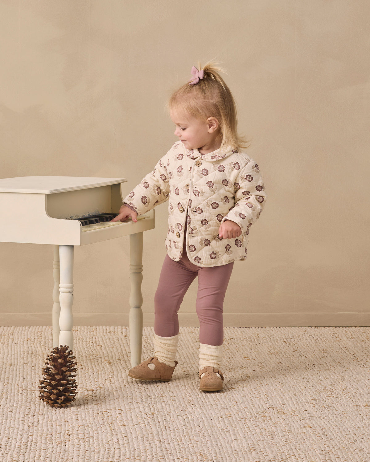 A toddler girl stands next to a small white table in a studio setting, wearing a cream-colored jacket with a brown pattern, mauve leggings, and brown shoes.
