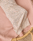 A close-up of a baby blanket with a small beige and blue floral print, draped over a textured dusty pink waffle-knit blanket on a woven rattan chair, with a baby's foot visible.
