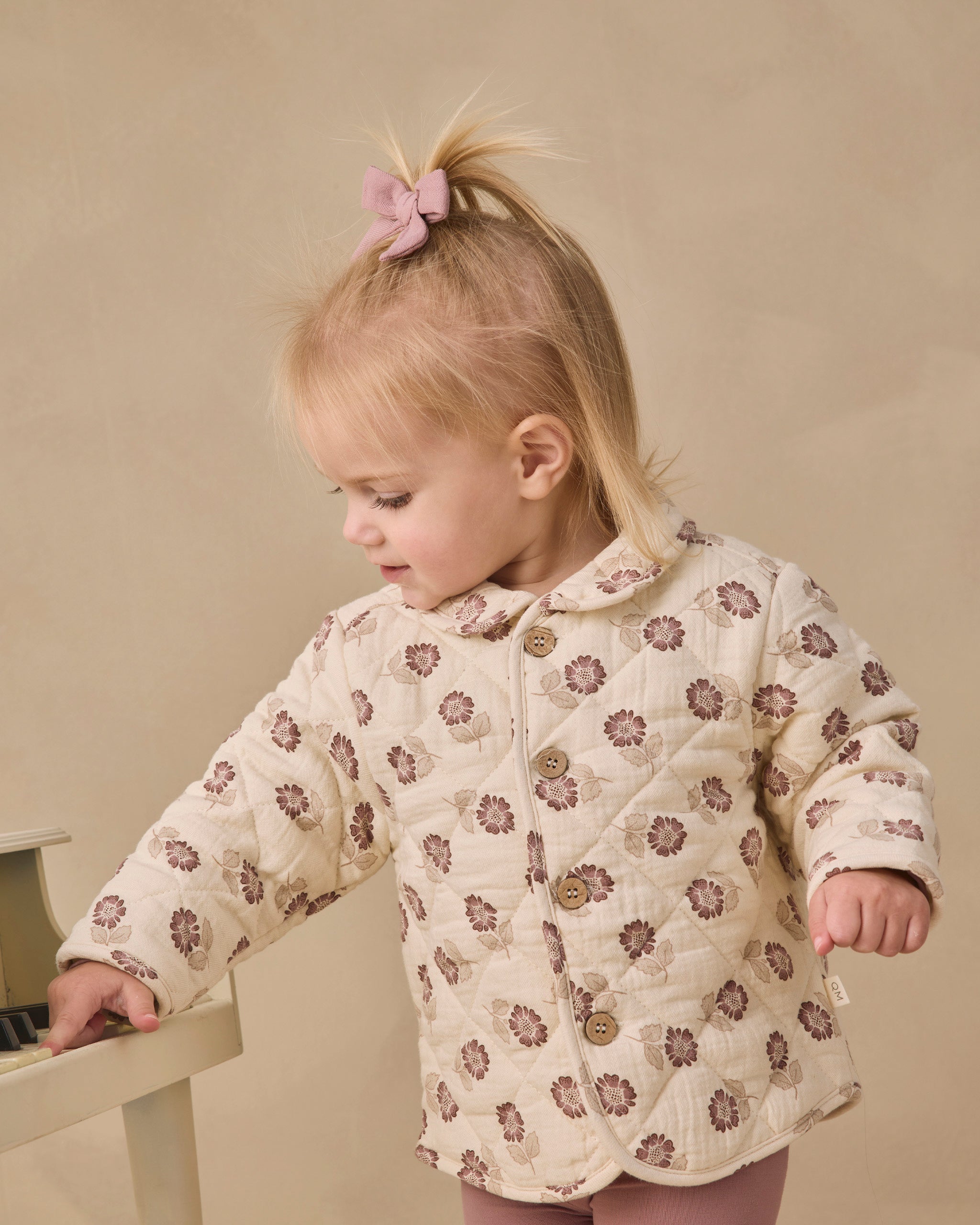 A toddler girl with blonde hair in a ponytail wearing a cream quilted jacket with a brown floral pattern, standing next to a small wooden table.