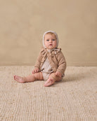 A baby sitting on a textured cream woven rug against a warm beige studio wall, wearing a beige knit bonnet and matching knit outfit.