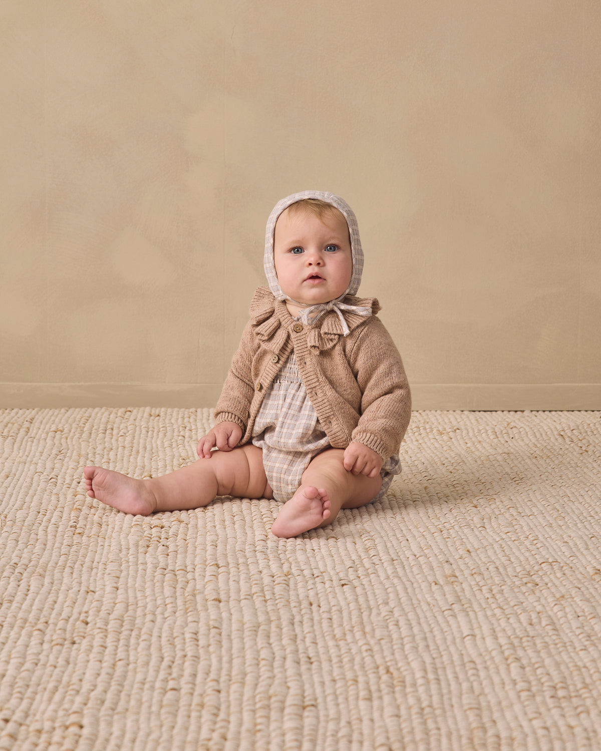 A baby sitting on a textured cream woven rug against a warm beige studio wall, wearing a beige knit bonnet and matching knit outfit.