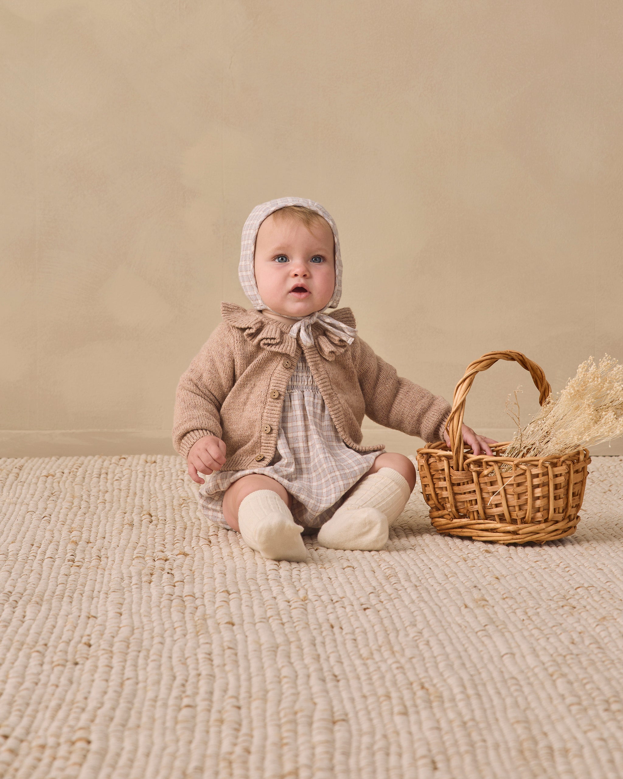 A baby wearing a beige knit bonnet, a brown ruffled cardigan, and a cream plaid romper sits on a textured cream rug next to a small wicker basket with dried grass.