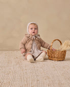 A baby wearing a beige knit bonnet, a brown ruffled cardigan, and a cream plaid romper sits on a textured cream rug next to a small wicker basket with dried grass.