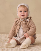 A baby sitting on a textured cream rug against a warm beige wall, wearing a beige knit bonnet, a brown cardigan, and cream socks, with a wicker basket to the side.