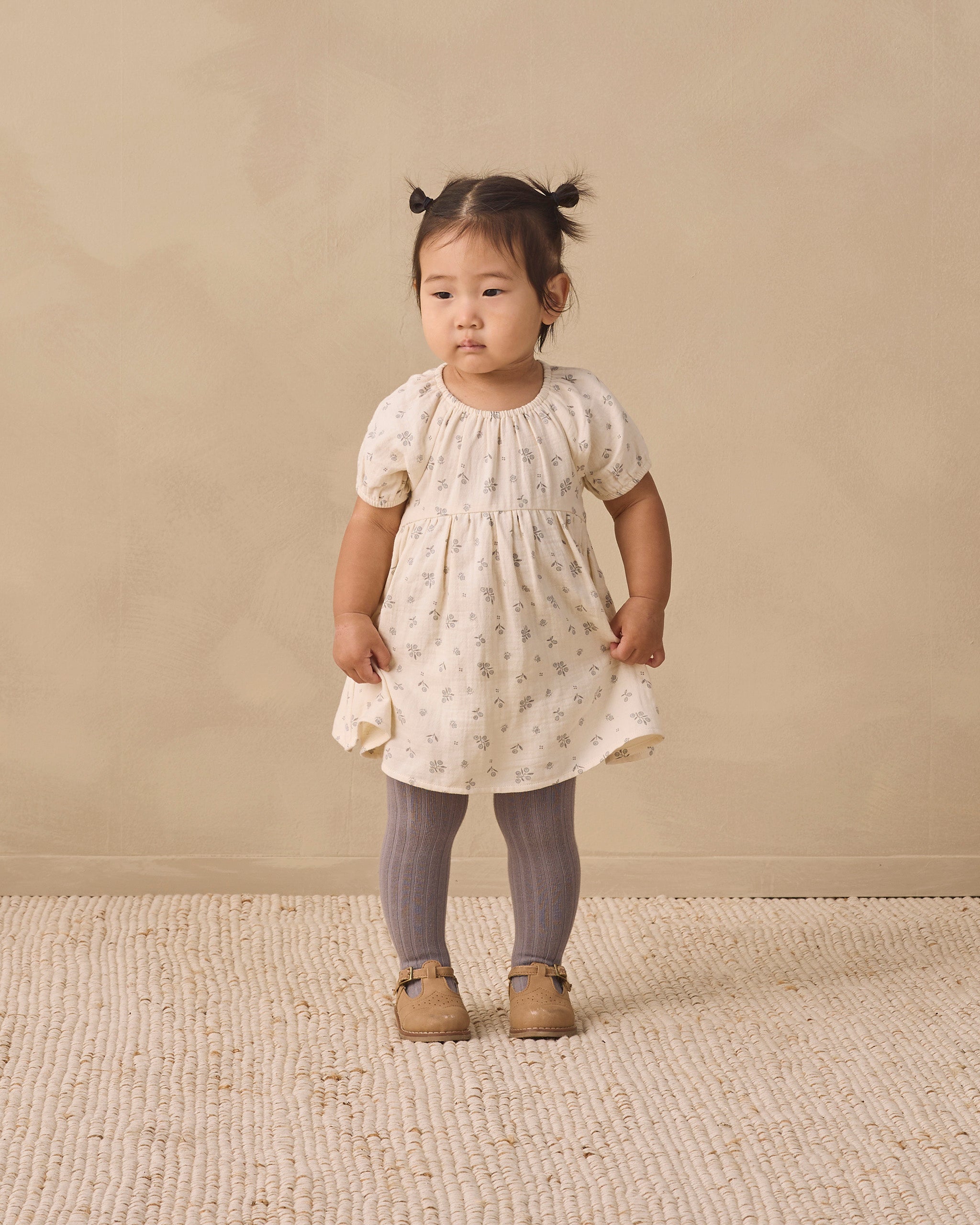 A toddler girl stands in a studio wearing a cream short-sleeve dress with a tiny floral print, smocked bodice, and ruffled hem, paired with gray tights and tan shoes, against a warm beige backdrop.