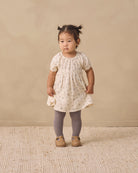 A toddler girl stands in a studio wearing a cream short-sleeve dress with a tiny floral print, smocked bodice, and ruffled hem, paired with gray tights and tan shoes, against a warm beige backdrop.
