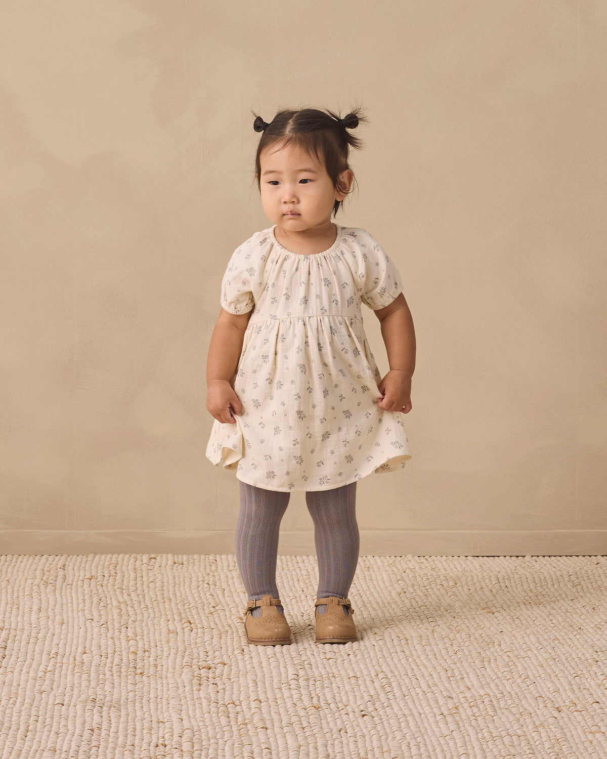 A toddler girl stands in a studio wearing a cream short-sleeve dress with a tiny floral print, smocked bodice, and ruffled hem, paired with gray tights and tan shoes, against a warm beige backdrop.