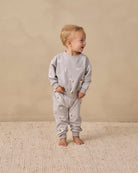 A toddler standing barefoot on a textured cream rug against a warm beige wall, wearing a light gray long-sleeve sweatshirt and matching sweatpants with a small celestial print of stars and crescent moons.