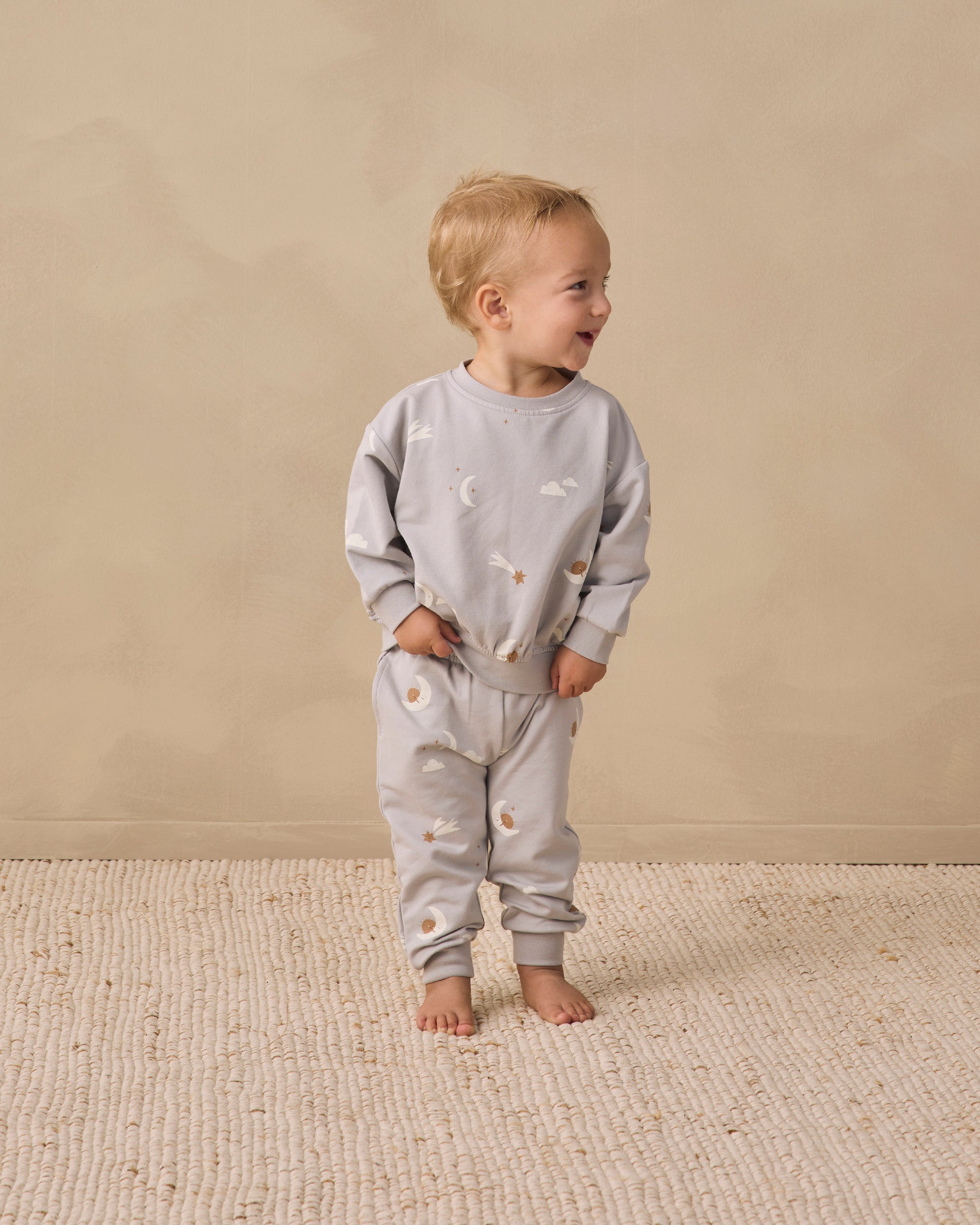 A toddler standing barefoot on a textured cream rug against a warm beige wall, wearing a light gray long-sleeve sweatshirt and matching sweatpants with a small celestial print of stars and crescent moons.