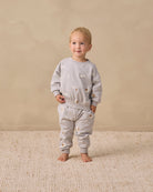A toddler stands barefoot on a textured cream rug against a warm beige studio wall, wearing a light gray long-sleeve sweatshirt and matching jogger pants with small scattered animal illustrations.