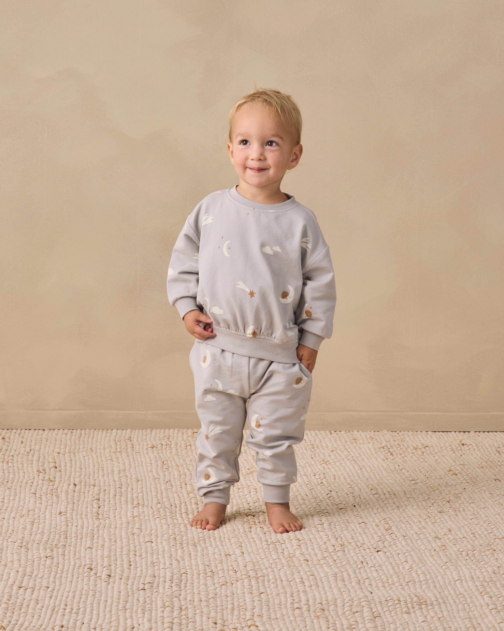 A toddler stands barefoot on a textured cream rug against a warm beige studio wall, wearing a light gray long-sleeve sweatshirt and matching jogger pants with small scattered animal illustrations.