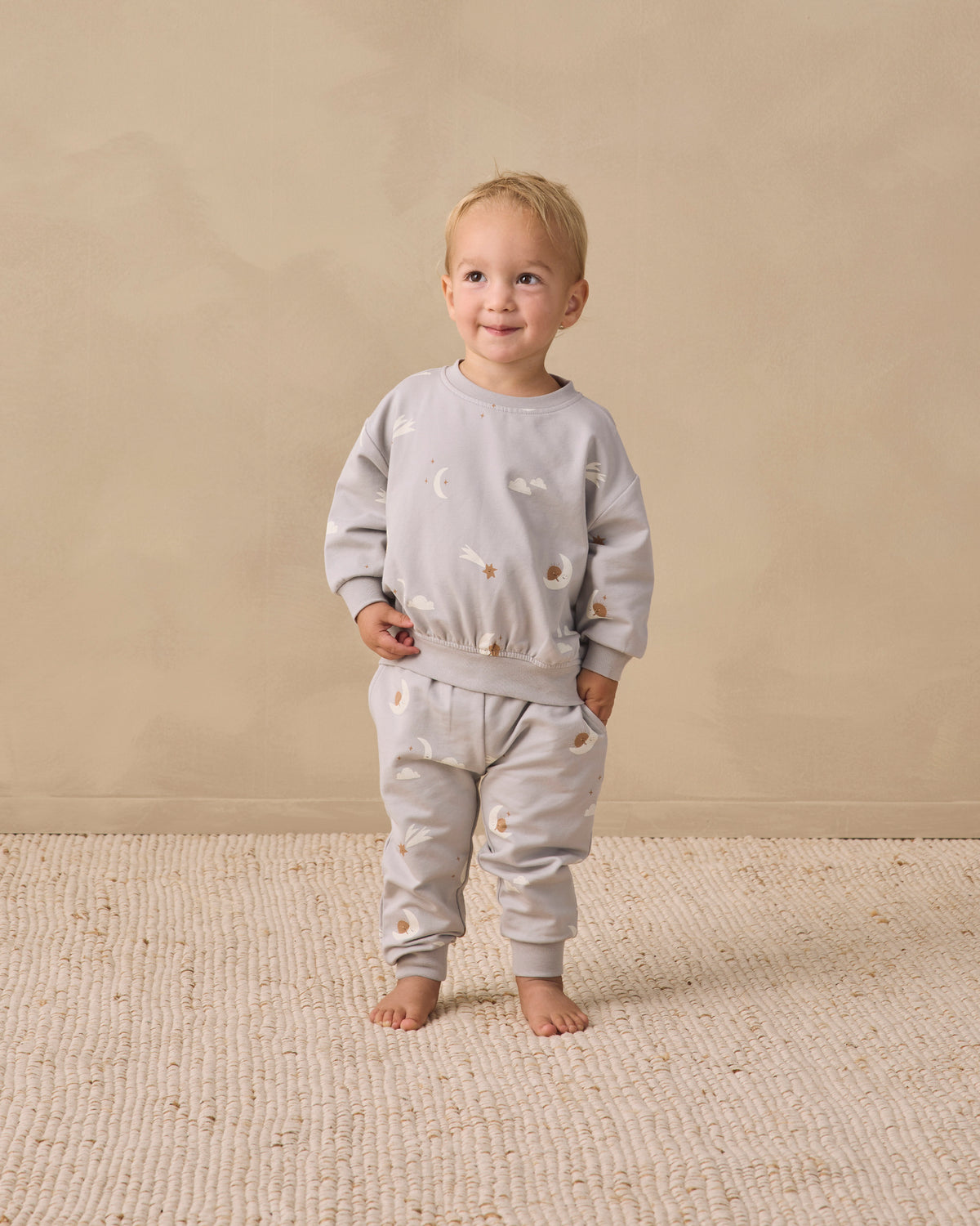 A toddler stands barefoot on a textured cream rug against a warm beige studio wall, wearing a light gray long-sleeve sweatshirt and matching jogger pants with small scattered animal illustrations.