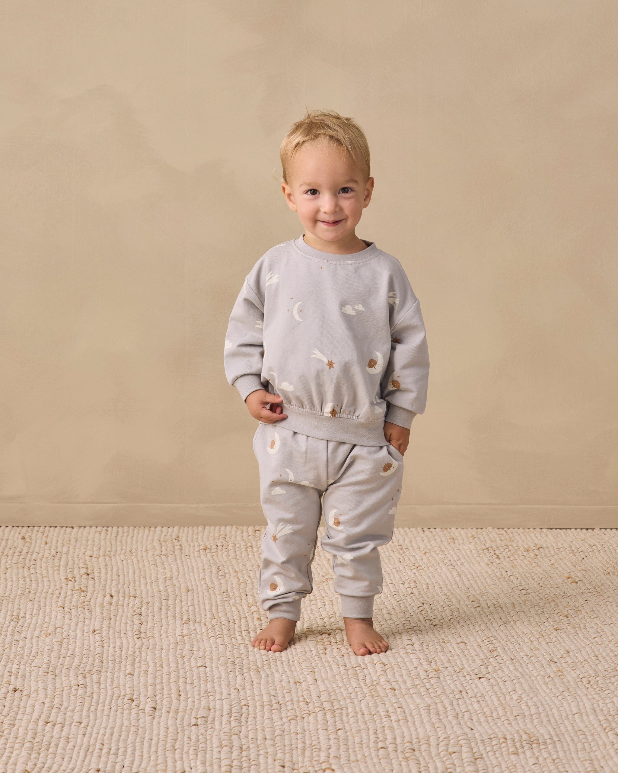 A toddler stands barefoot on a textured cream rug against a warm beige wall, wearing a matching light gray sweatshirt and jogger pants set with small embroidered details.