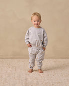 A toddler stands barefoot on a textured cream rug against a warm beige wall, wearing a matching light gray sweatshirt and jogger pants set with small embroidered details.