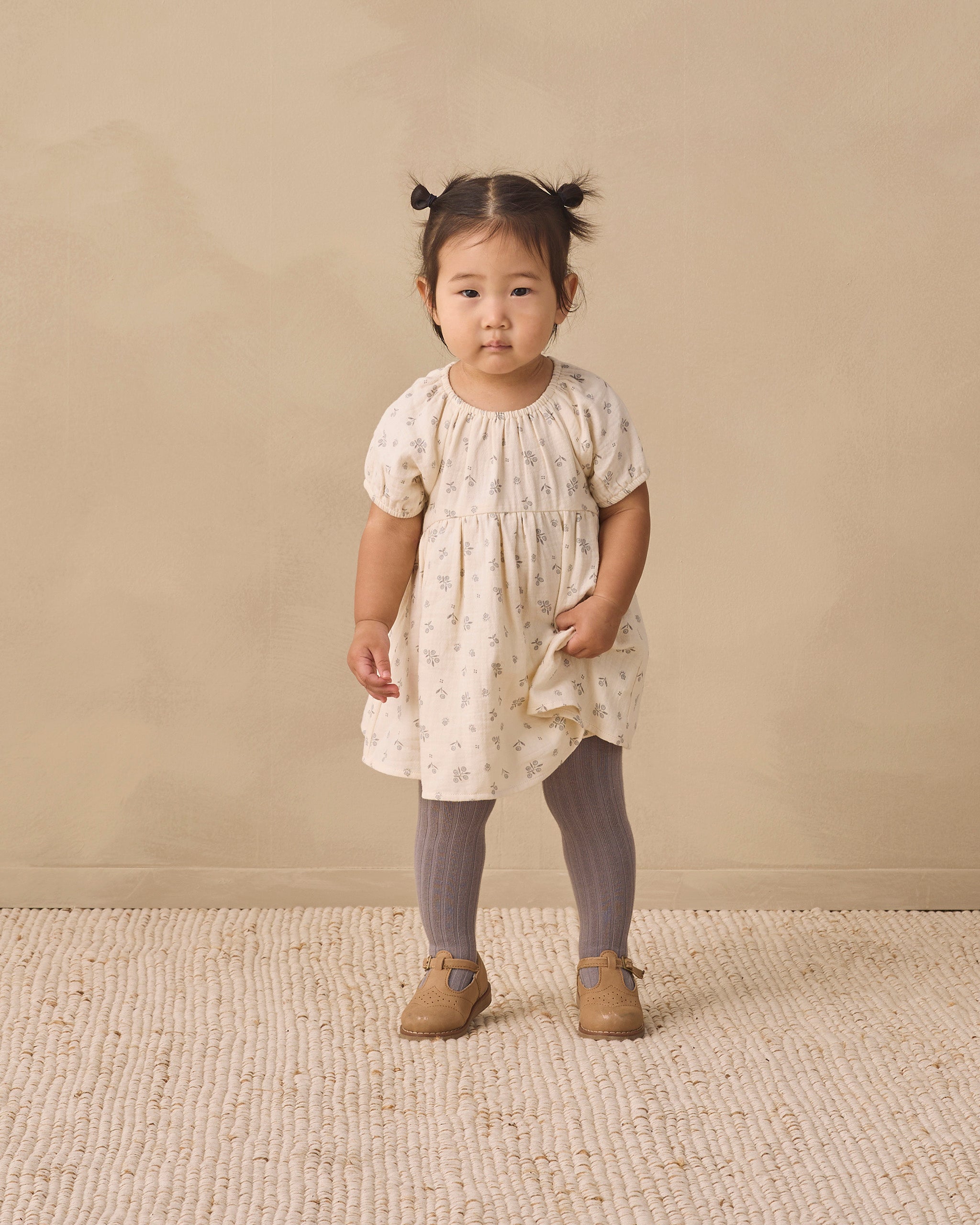 A toddler girl stands in a studio wearing a cream short-sleeve dress with a small beige floral print, gray tights, and tan shoes against a warm beige backdrop.