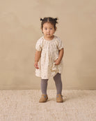 A toddler girl stands in a studio wearing a cream short-sleeve dress with a small beige floral print, gray tights, and tan shoes against a warm beige backdrop.